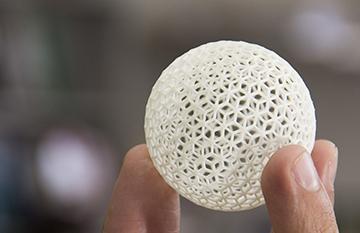 A hand holding a small white sphere with an intricate 3D-printed lattice.