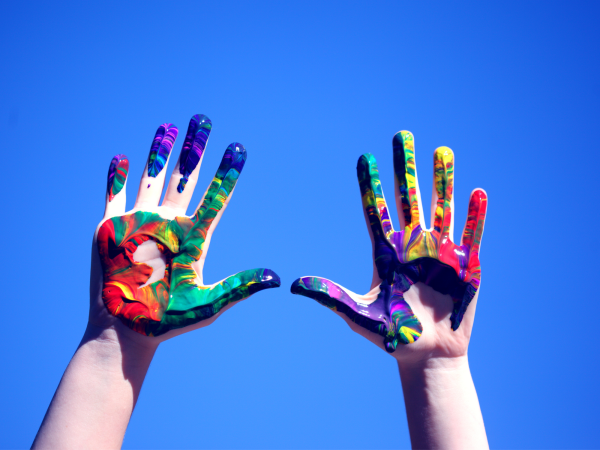 Two raised hands covered in bright paint against a blue background.
