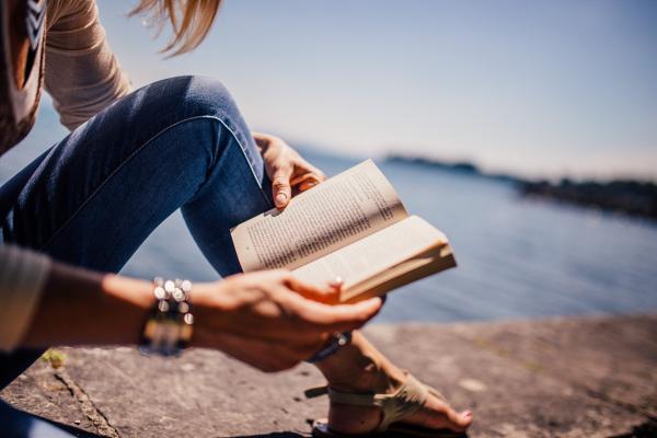 A person reading an open book outdoors beside the water.
