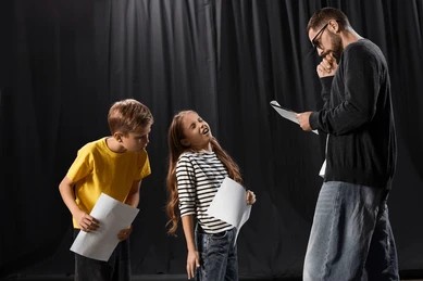 Two children and an adult holding scripts beside a black stage curtain.